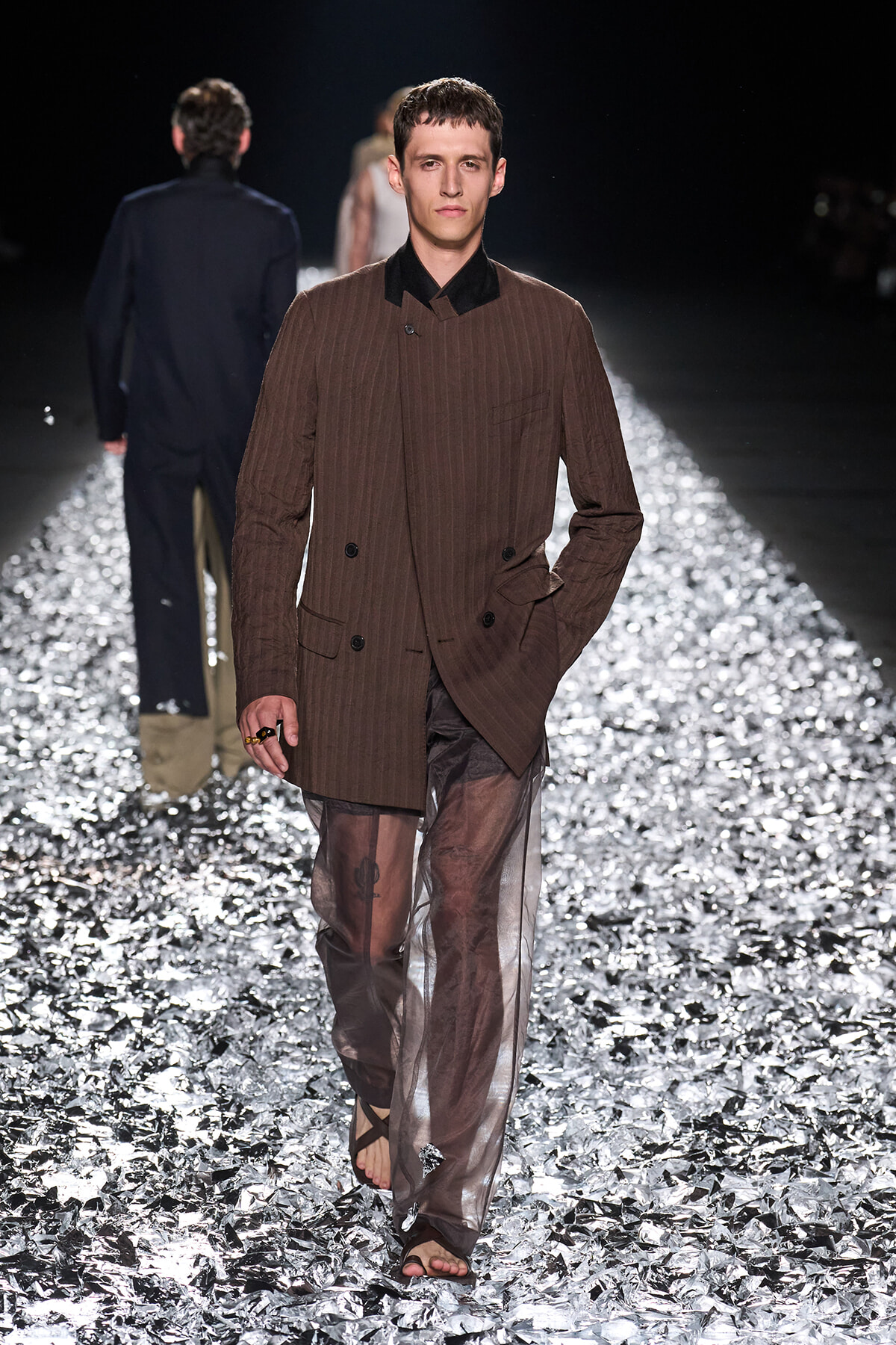 Male model walks a fashion runway in a brown pinstriped double-breasted blazer and sheer brown pants, barefoot sandals on a metallic foil floor.