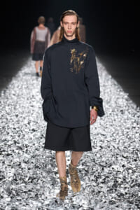 Male model walking a dark, oversized navy top with gold embroidery on the chest, black shorts, snakeskin boots on a reflective runway surface.