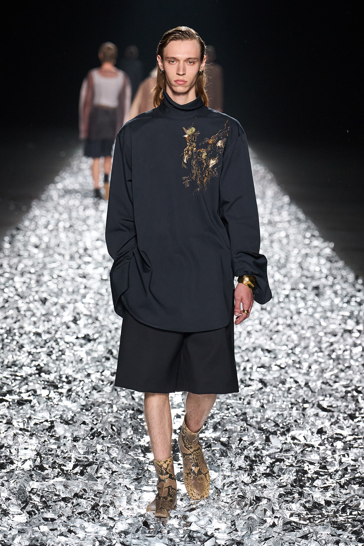 Male model walking a dark, oversized navy top with gold embroidery on the chest, black shorts, snakeskin boots on a reflective runway surface.