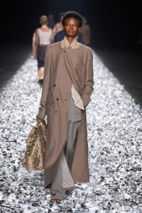 Model walks a fashion runway wearing a long beige trench over taupe and gray layers, carrying a snakeskin bag.