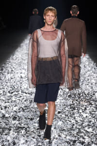 Male model walking a fashion runway on a reflective foil floor, wearing a sheer brown overlay top over a white tank and navy shorts with a patterned belt.