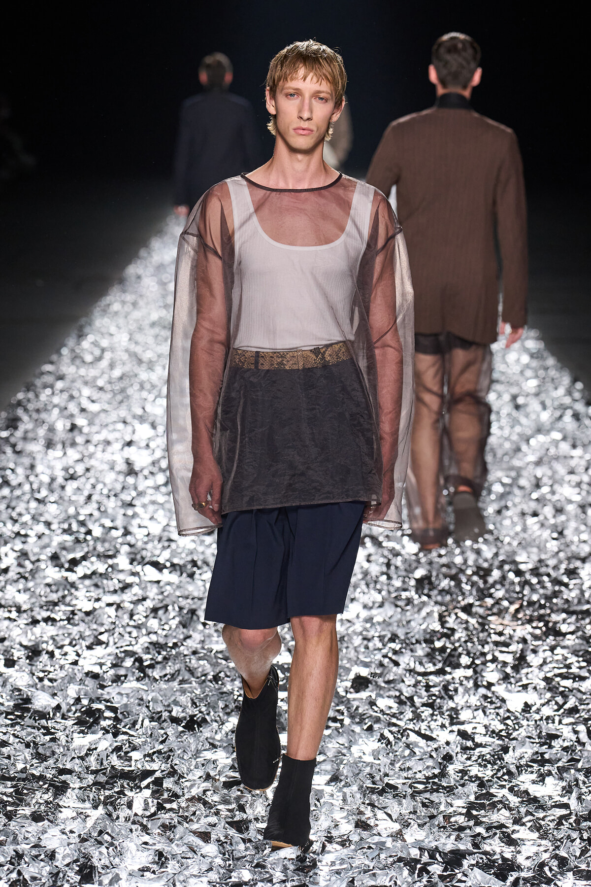 Male model walking a fashion runway on a reflective foil floor, wearing a sheer brown overlay top over a white tank and navy shorts with a patterned belt.