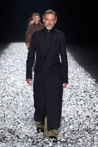 Male model in a long navy double‑breasted coat over beige wide‑leg trousers walks a glittery runway, beard gray.