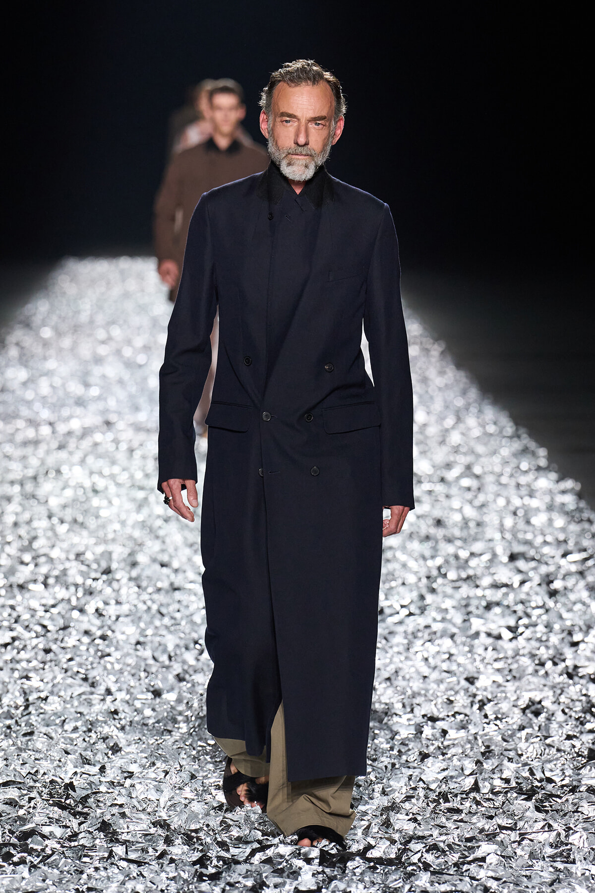 Male model in a long navy double‑breasted coat over beige wide‑leg trousers walks a glittery runway, beard gray.