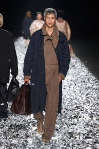 Male model walking a runway in a navy overcoat over brown utilitarian jumpsuit, carrying a brown leather duffel bag.