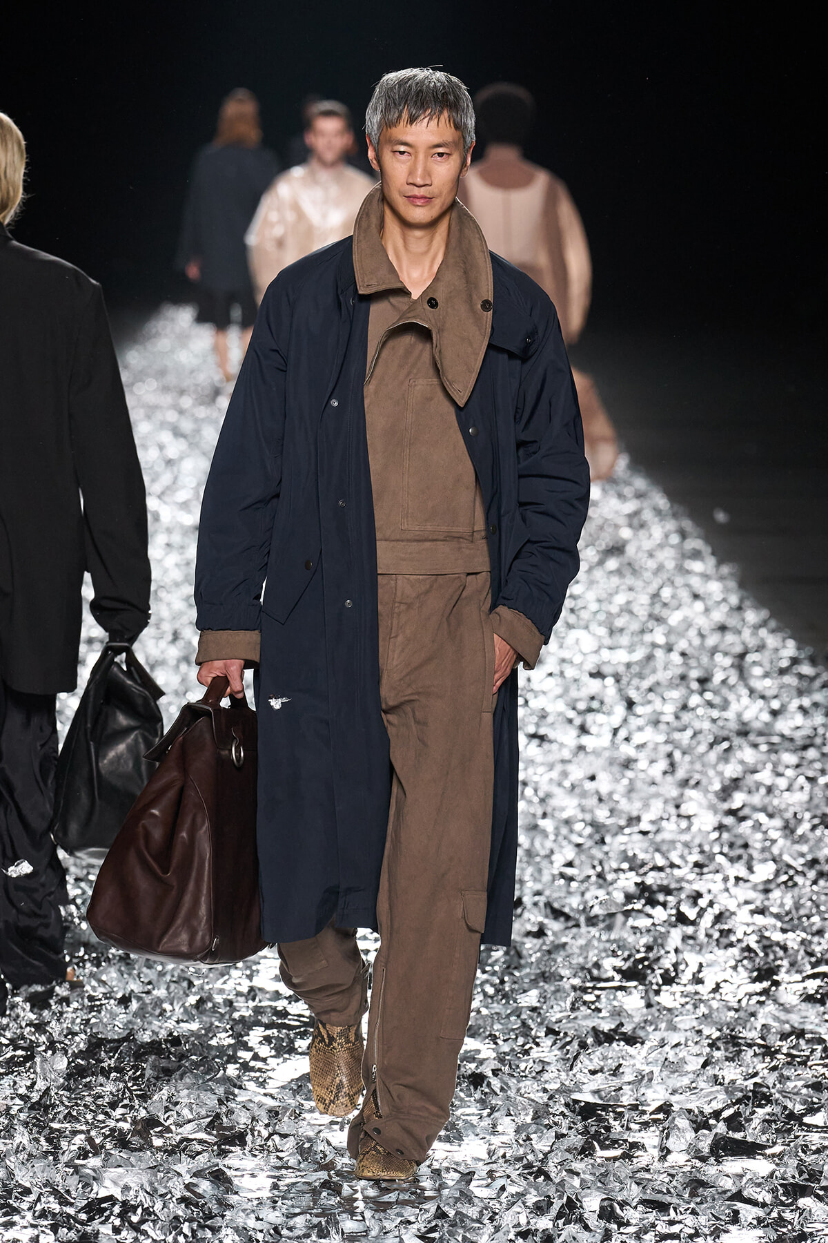Male model walking a runway in a navy overcoat over brown utilitarian jumpsuit, carrying a brown leather duffel bag.