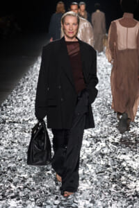 Model walks a fashion runway in an oversized black blazer, burgundy top, and wide black pants, carrying a black leather bag on a glittery silver runway.
