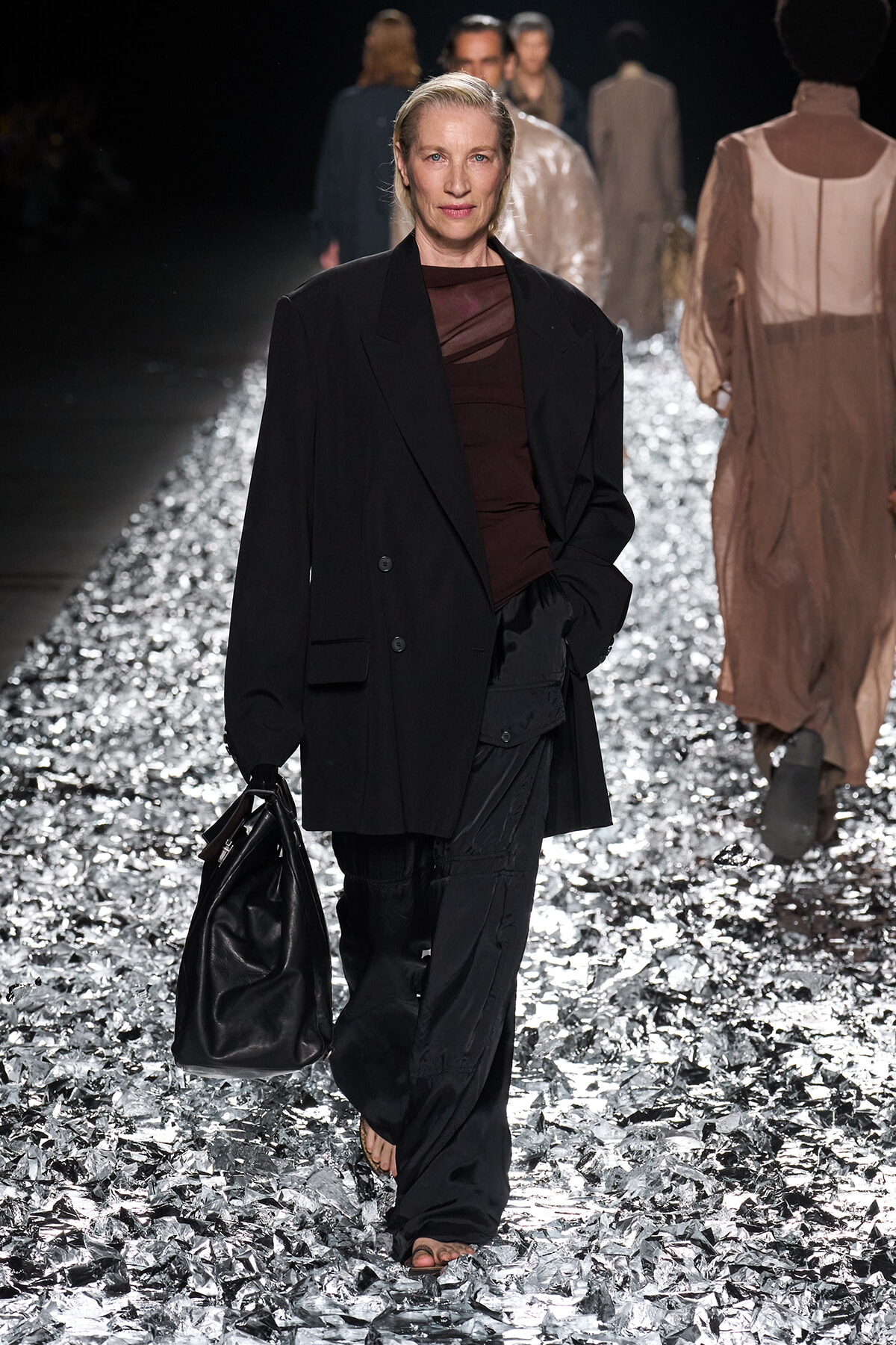 Model walks a fashion runway in an oversized black blazer, burgundy top, and wide black pants, carrying a black leather bag on a glittery silver runway.