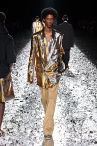 Model walking a runway in a metallic gold blazer and tan pants on a glittering catwalk.