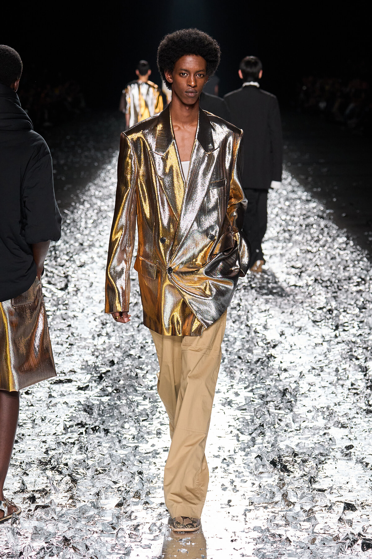 Model walking a runway in a metallic gold blazer and tan pants on a glittering catwalk.