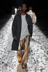 Male model walking a fashion runway in a black coat over a silver sparkly sweater and metallic gold pants on a glimmering reflective catwalk.