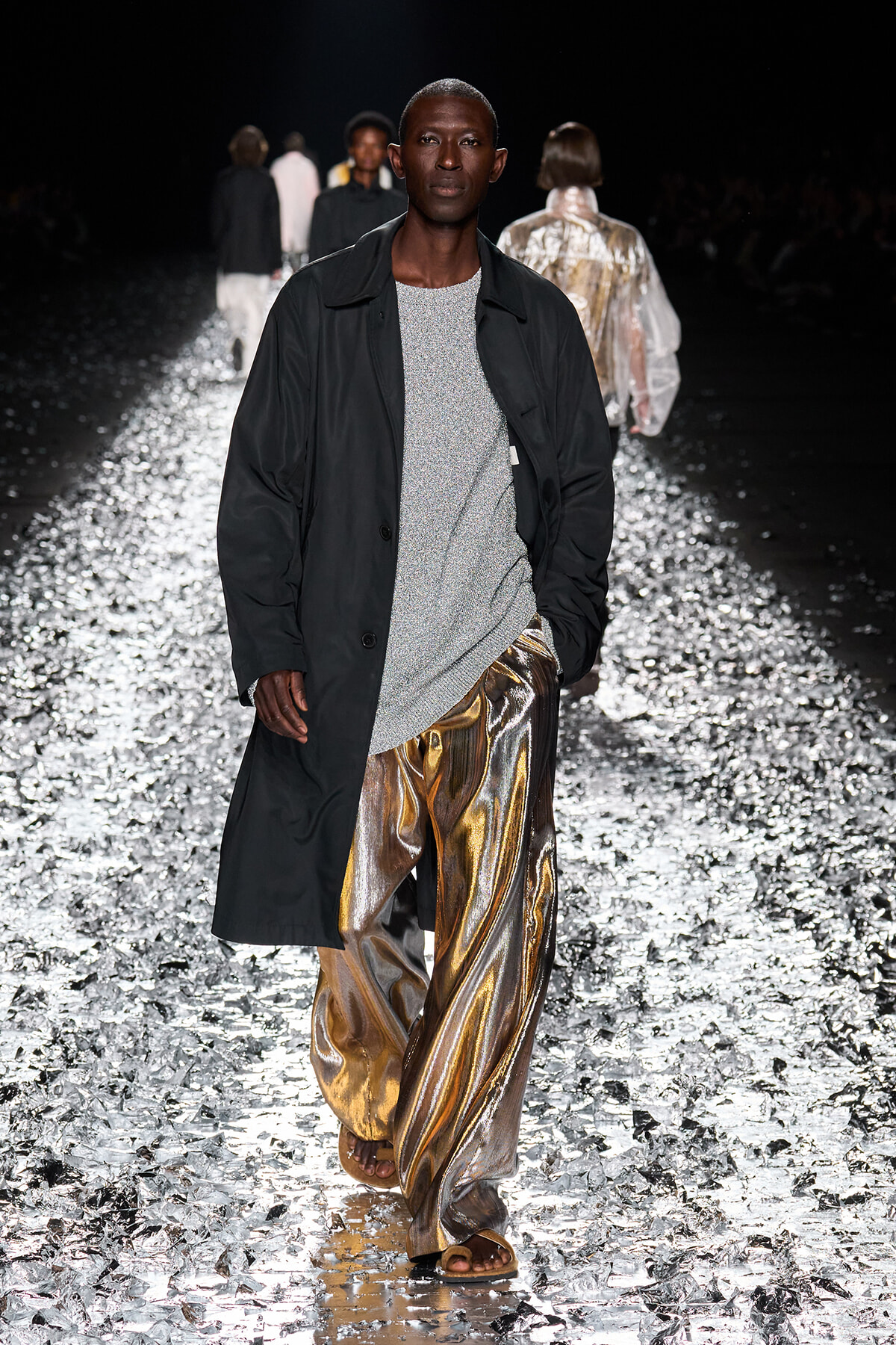 Male model walking a fashion runway in a black coat over a silver sparkly sweater and metallic gold pants on a glimmering reflective catwalk.
