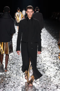Male fashion model walking a runway in a long black coat over metallic gold pants and sandals.