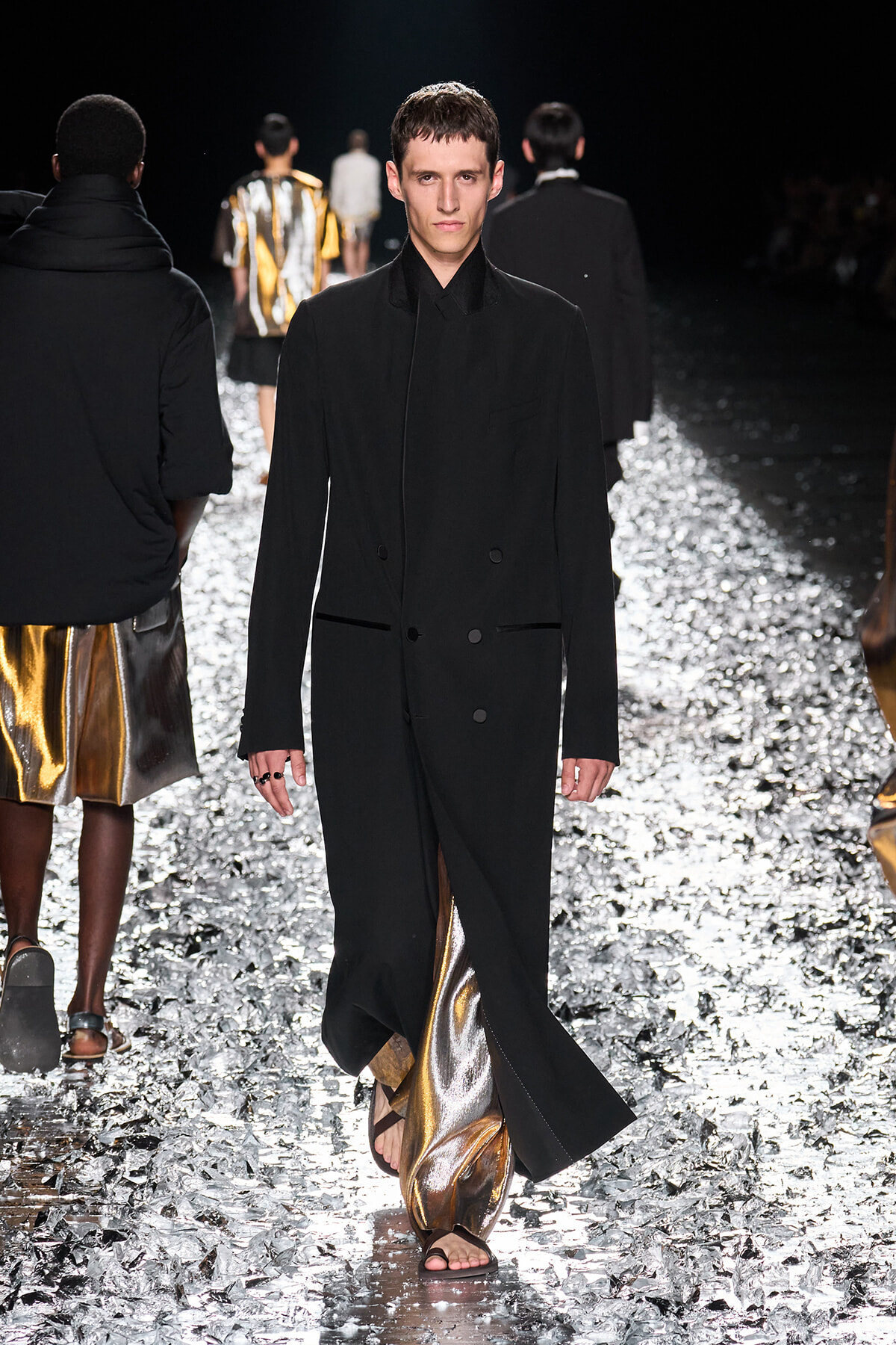 Male fashion model walking a runway in a long black coat over metallic gold pants and sandals.