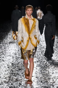 Male model walks a fashion runway in a gold-and-cream brocade blazer, beige top, metallic gold shorts, and brown fur slides on a glittering runway surface, dark backdrop.