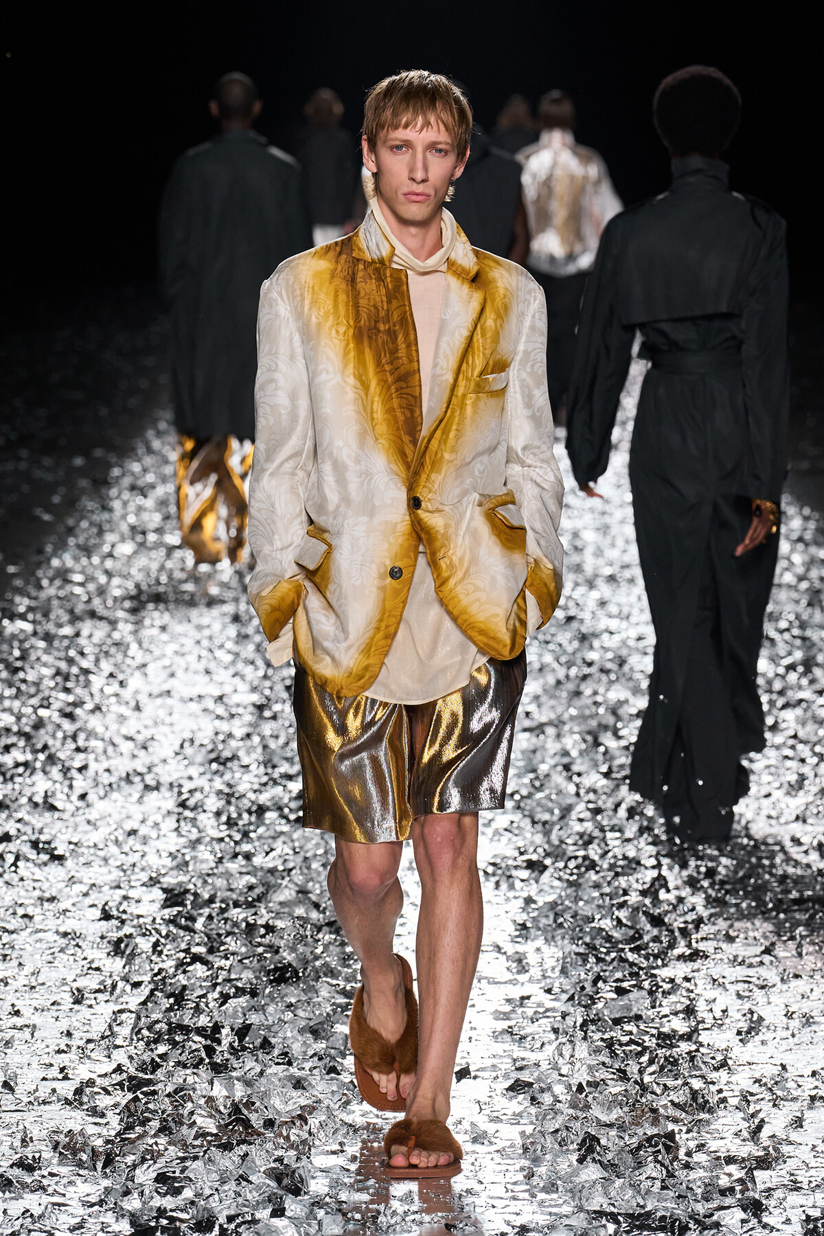 Male model walks a fashion runway in a gold-and-cream brocade blazer, beige top, metallic gold shorts, and brown fur slides on a glittering runway surface, dark backdrop.