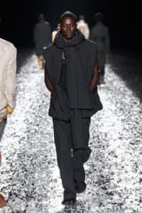 Male model in oversized black outerwear and scarf walks a glittery runway at a fashion show.