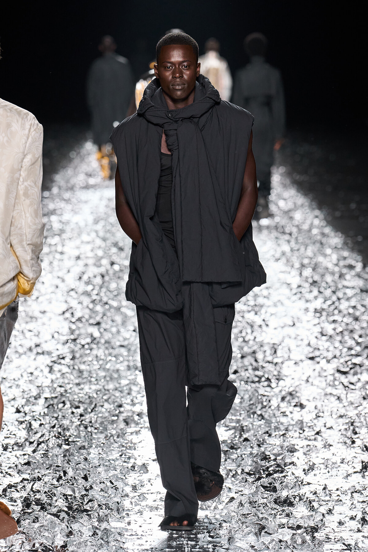 Male model in oversized black outerwear and scarf walks a glittery runway at a fashion show.