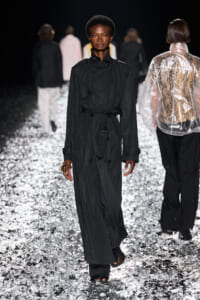 Female model walks a fashion runway in a long black trench coat belted at the waist, on a reflective, silver-crystal floor with blurred background models behind her.