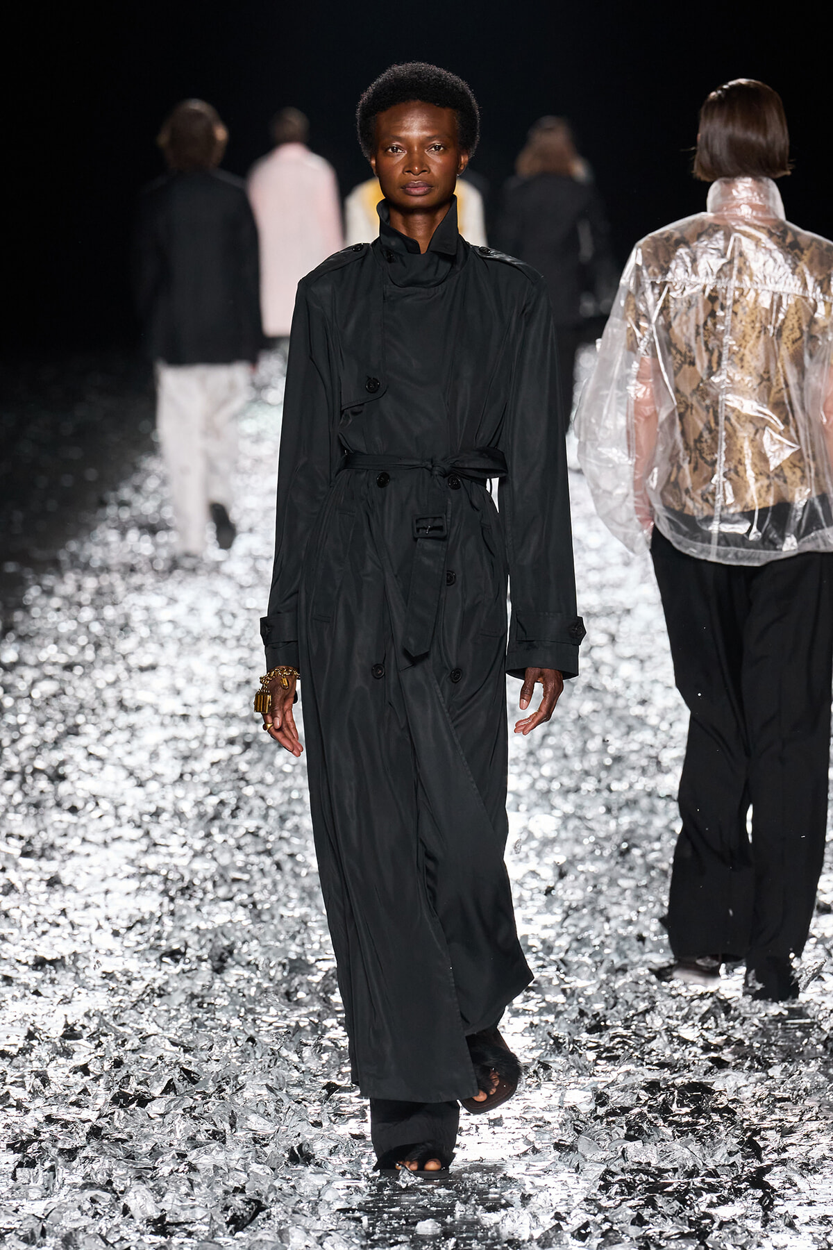 Female model walks a fashion runway in a long black trench coat belted at the waist, on a reflective, silver-crystal floor with blurred background models behind her.