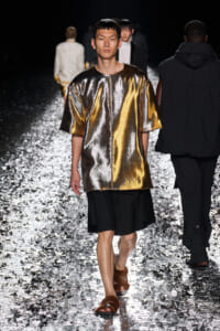 Male model walks the runway in a metallic gold oversized top with black shorts and brown fur sandals on a reflective, glittering catwalk.