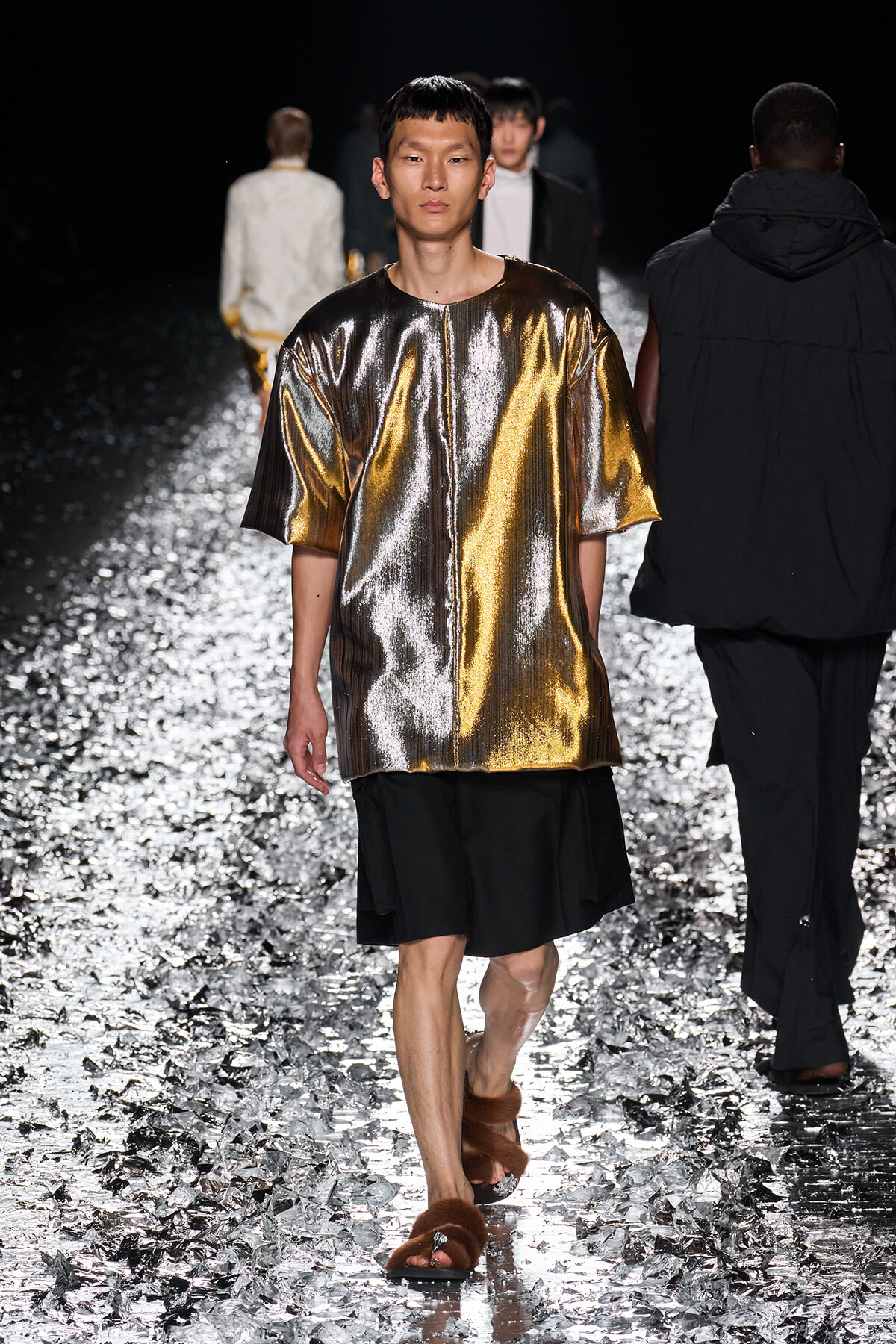 Male model walks the runway in a metallic gold oversized top with black shorts and brown fur sandals on a reflective, glittering catwalk.