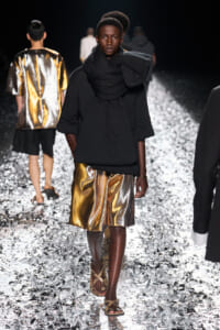 Model walks a fashion runway in a black oversized top with a large wrapped scarf, metallic gold shorts, and strappy sandals; confetti-covered catwalk.