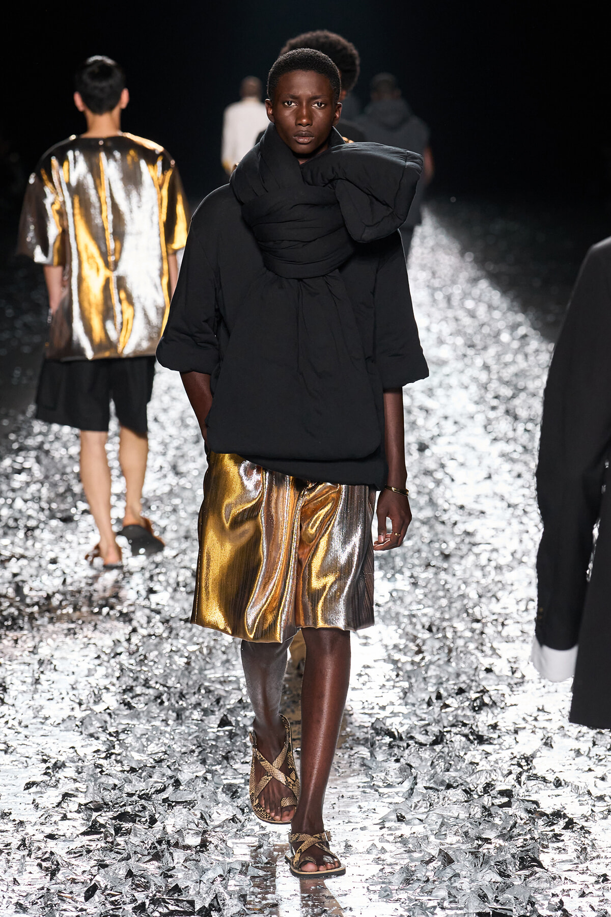 Model walks a fashion runway in a black oversized top with a large wrapped scarf, metallic gold shorts, and strappy sandals; confetti-covered catwalk.