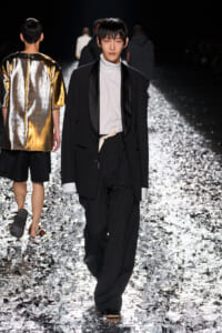 Male model walks the runway in a black suit with a white turtleneck and a beige belt, on a glittering, reflective runway surface.