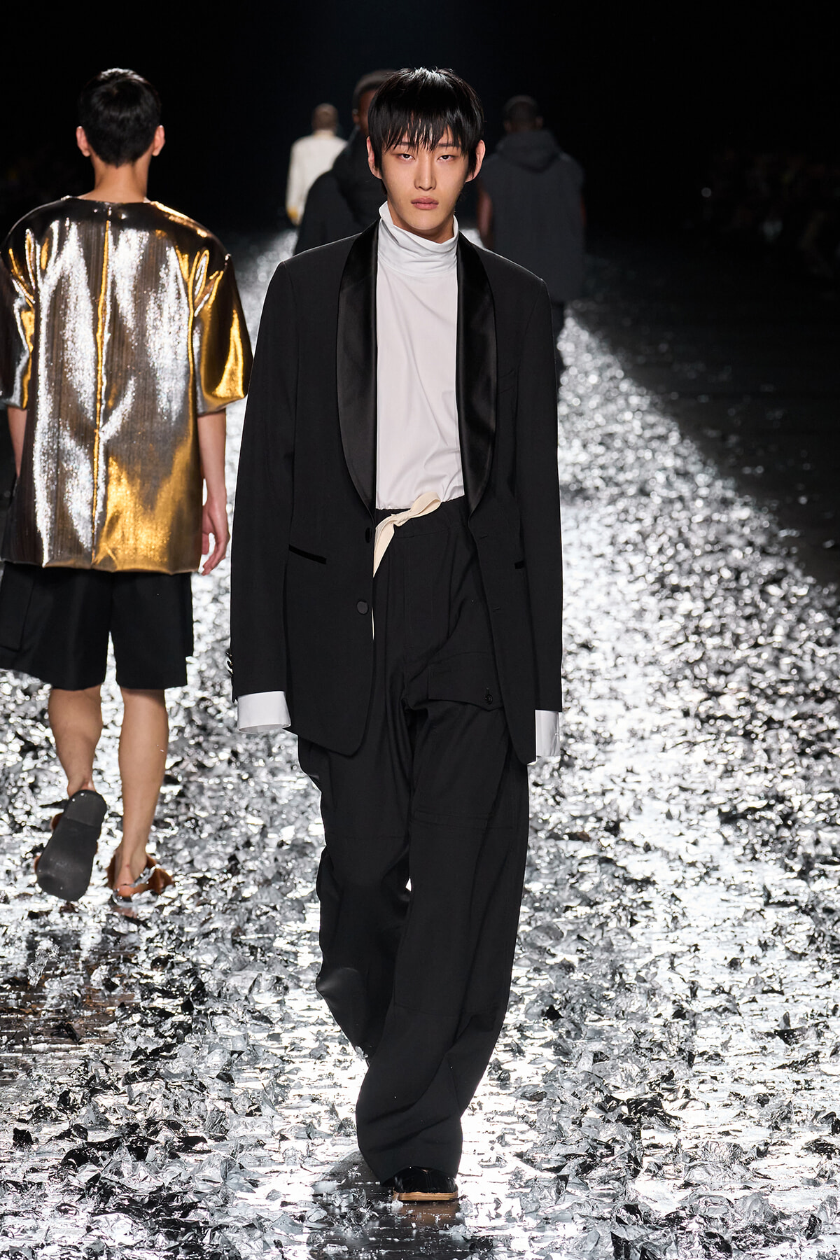 Male model walks the runway in a black suit with a white turtleneck and a beige belt, on a glittering, reflective runway surface.