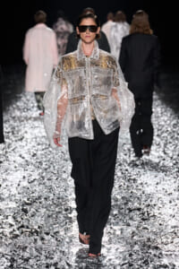 Model walks a runway in a oversized clear plastic jacket over a snakeskin top and black pants, wearing dark sunglasses on a glittering runway floor.