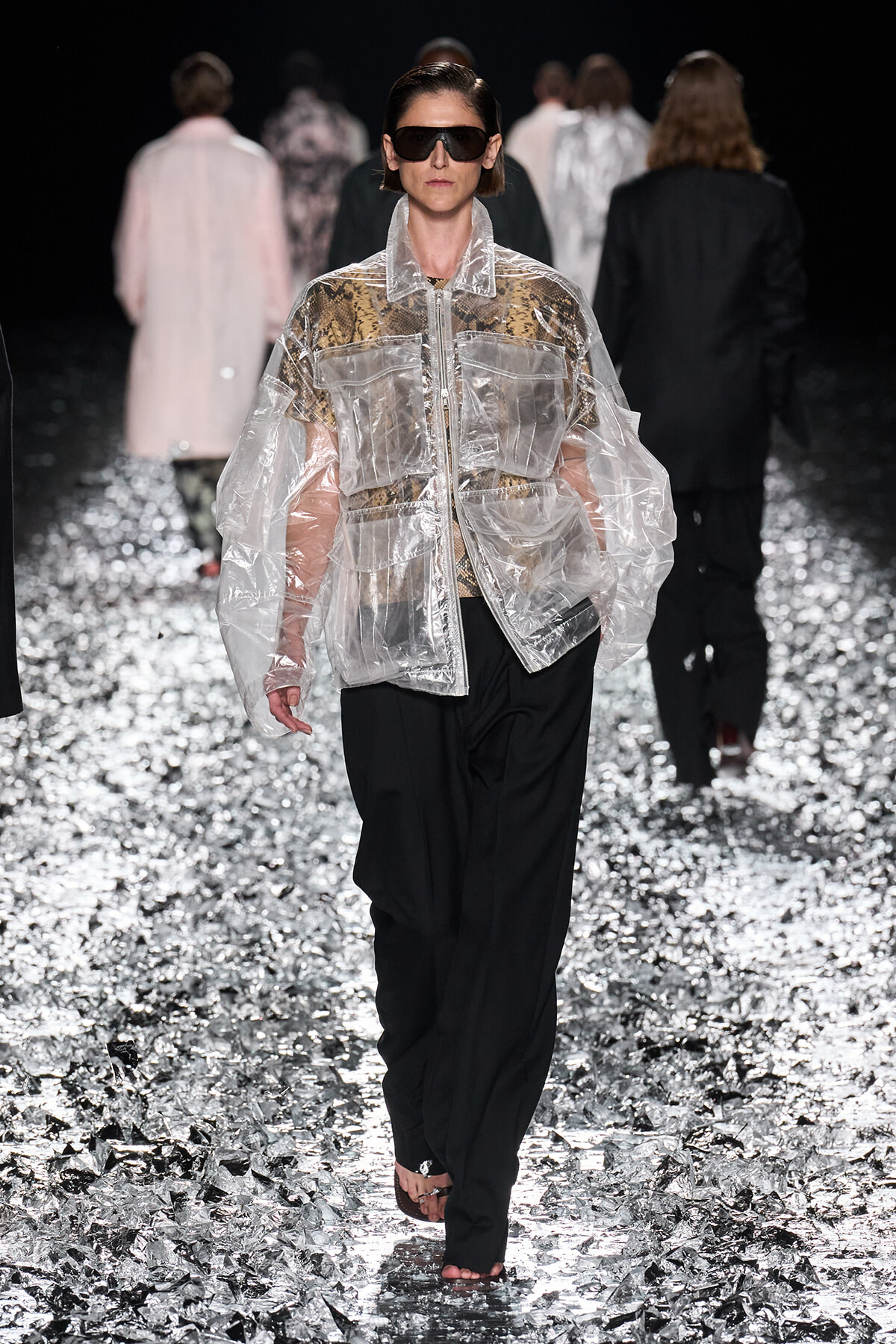Model walks a runway in a oversized clear plastic jacket over a snakeskin top and black pants, wearing dark sunglasses on a glittering runway floor.