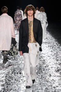 Model walks a runway in a black blazer over a snake-print top and white embroidered cargo pants, stepping through a sparkling reflective floor.
