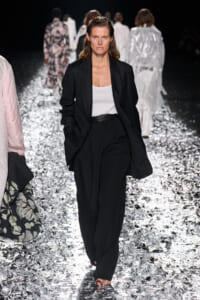 Model walking a fashion runway in a black blazer and wide-leg pants with a white tank top; glittering floor and other models in the background.
