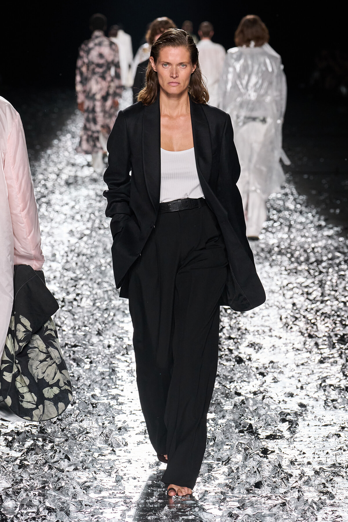 Model walking a fashion runway in a black blazer and wide-leg pants with a white tank top; glittering floor and other models in the background.