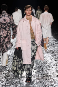 Male model on a fashion runway wearing a light pink quilted coat over a snake-print top and oversized floral-print pants, carrying a black bag.