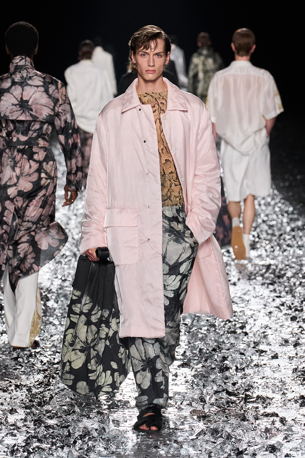 Male model on a fashion runway wearing a light pink quilted coat over a snake-print top and oversized floral-print pants, carrying a black bag.