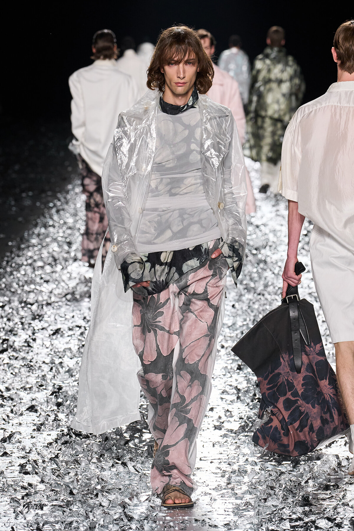 Male model on a fashion runway wearing a translucent raincoat over a gray top and pink floral-printed pants, walking on a glittery silver runway.