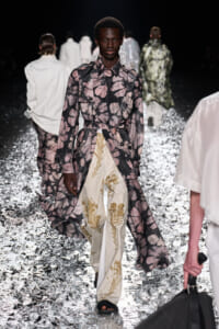 Model walks the runway in a long floral-print trench coat tied at the waist over cream floral trousers on a glittery, metallic runway surface.