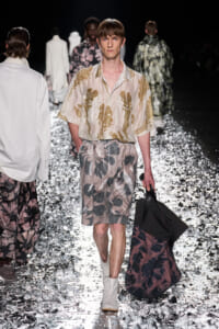 Male model on a fashion runway wearing beige floral shirt and matching shorts, carrying a large dark tote bag on a confetti-covered catwalk.