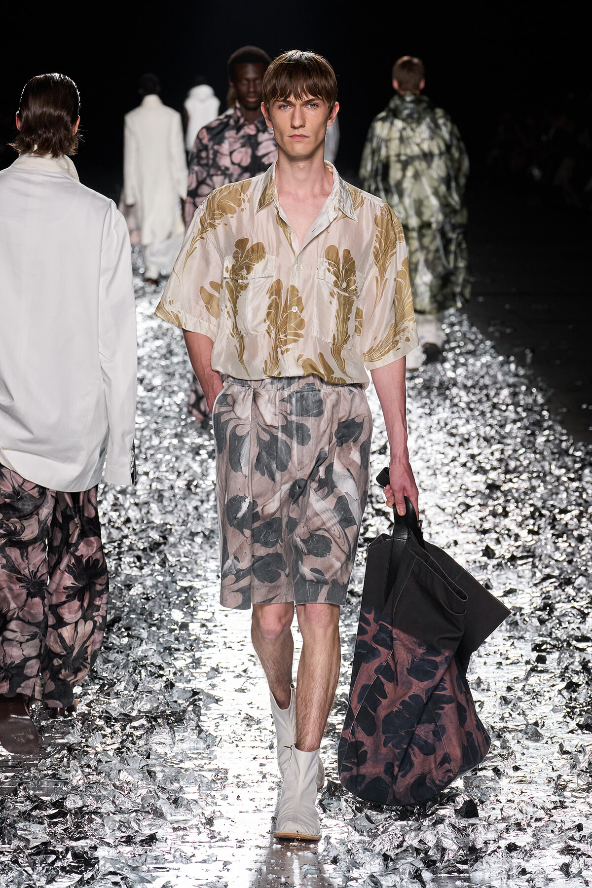 Male model on a fashion runway wearing beige floral shirt and matching shorts, carrying a large dark tote bag on a confetti-covered catwalk.