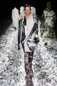 Model on a fashion show runway wearing a reflective silver oversized blazer, white shirt, and pink floral pants among silver confetti.