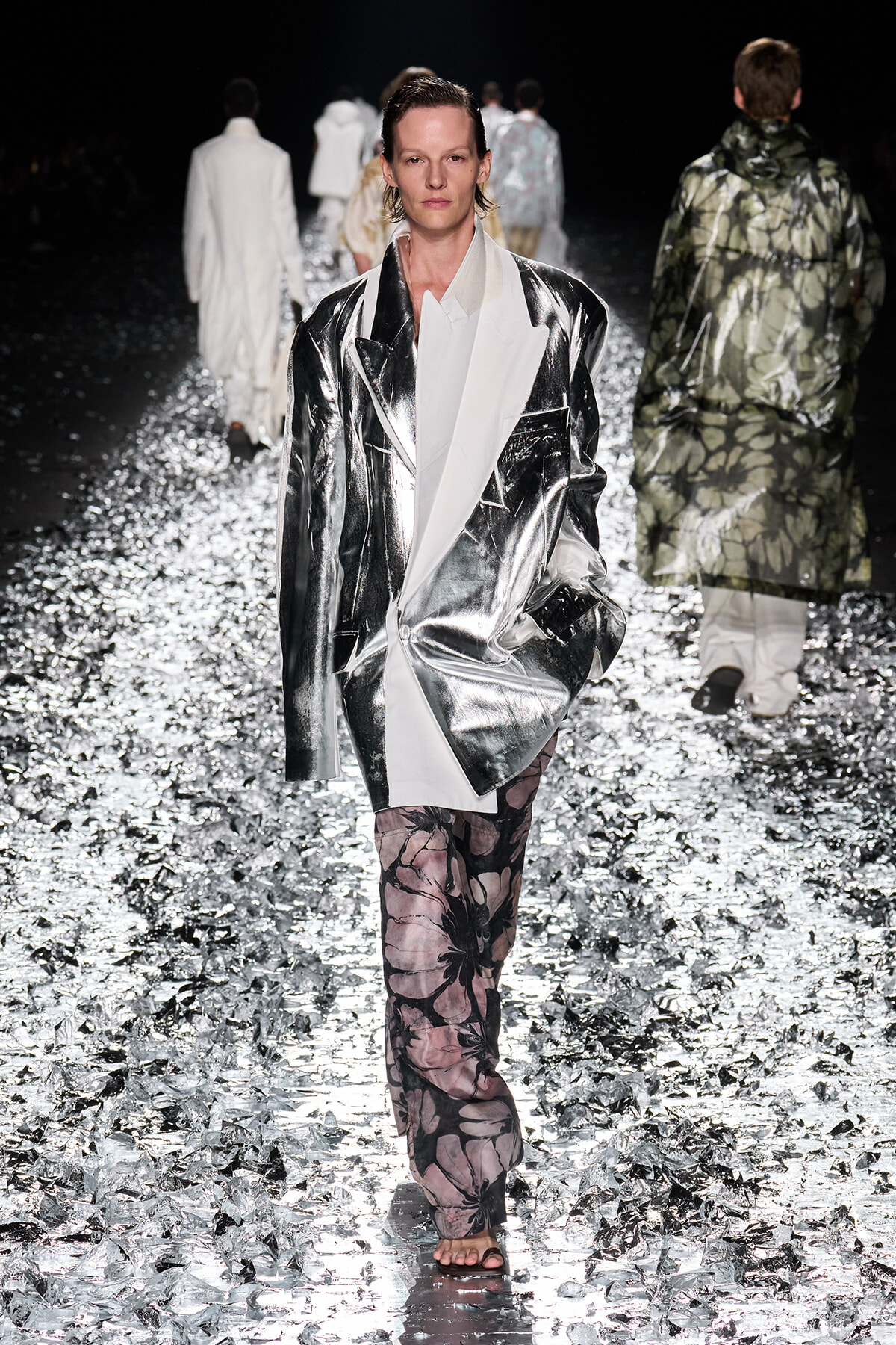 Model on a fashion show runway wearing a reflective silver oversized blazer, white shirt, and pink floral pants among silver confetti.