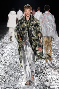 Male model walks a fashion runway in a glossy green floral raincoat over metallic silver wide-leg pants, surrounded by a glittering floor runway show setting.