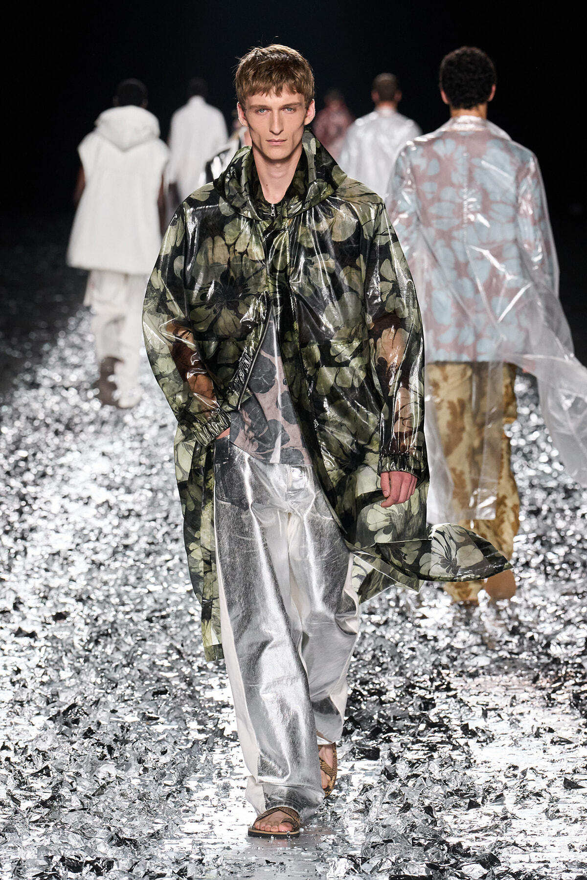 Male model walks a fashion runway in a glossy green floral raincoat over metallic silver wide-leg pants, surrounded by a glittering floor runway show setting.