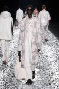 Model on a runway wearing a long pastel floral coat, oversized cream outfit, dark sunglasses, and carrying a white bag; metallic confetti on the floor add shine.
