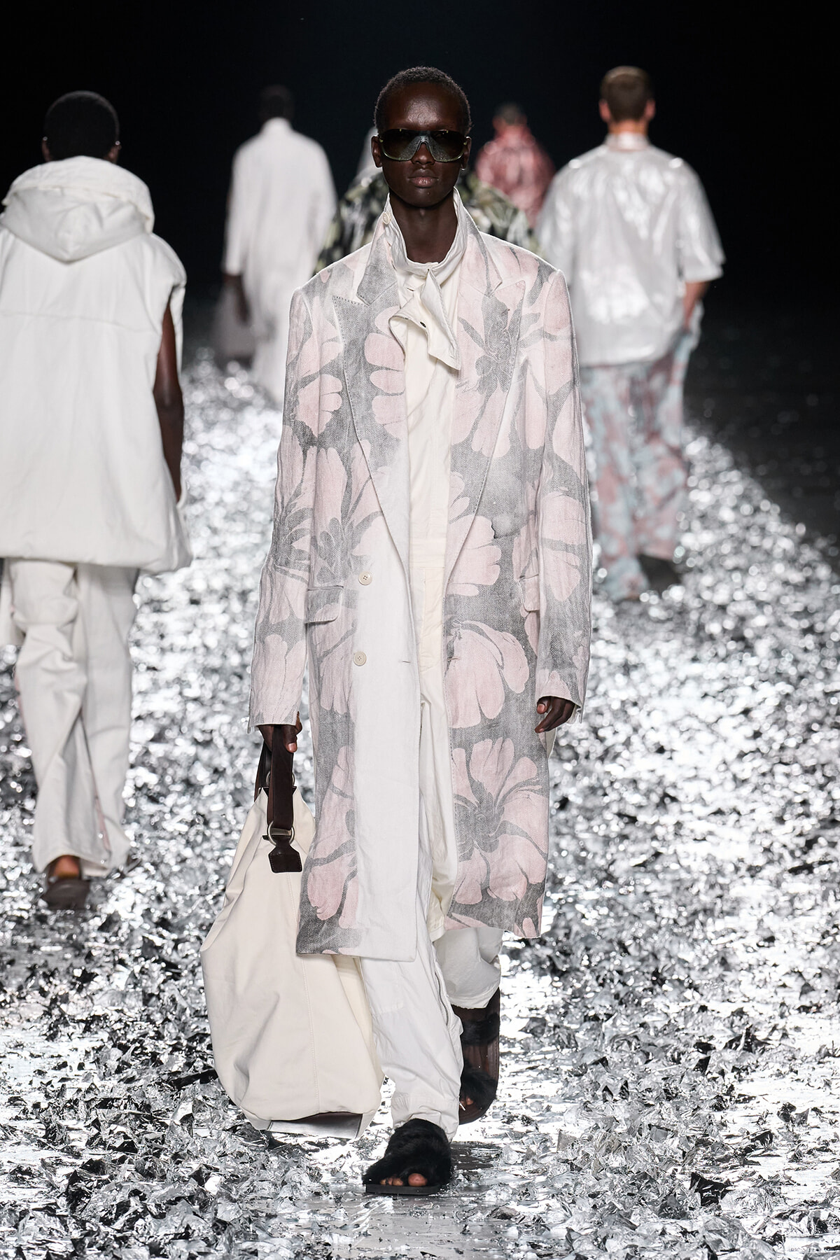 Model on a runway wearing a long pastel floral coat, oversized cream outfit, dark sunglasses, and carrying a white bag; metallic confetti on the floor add shine.