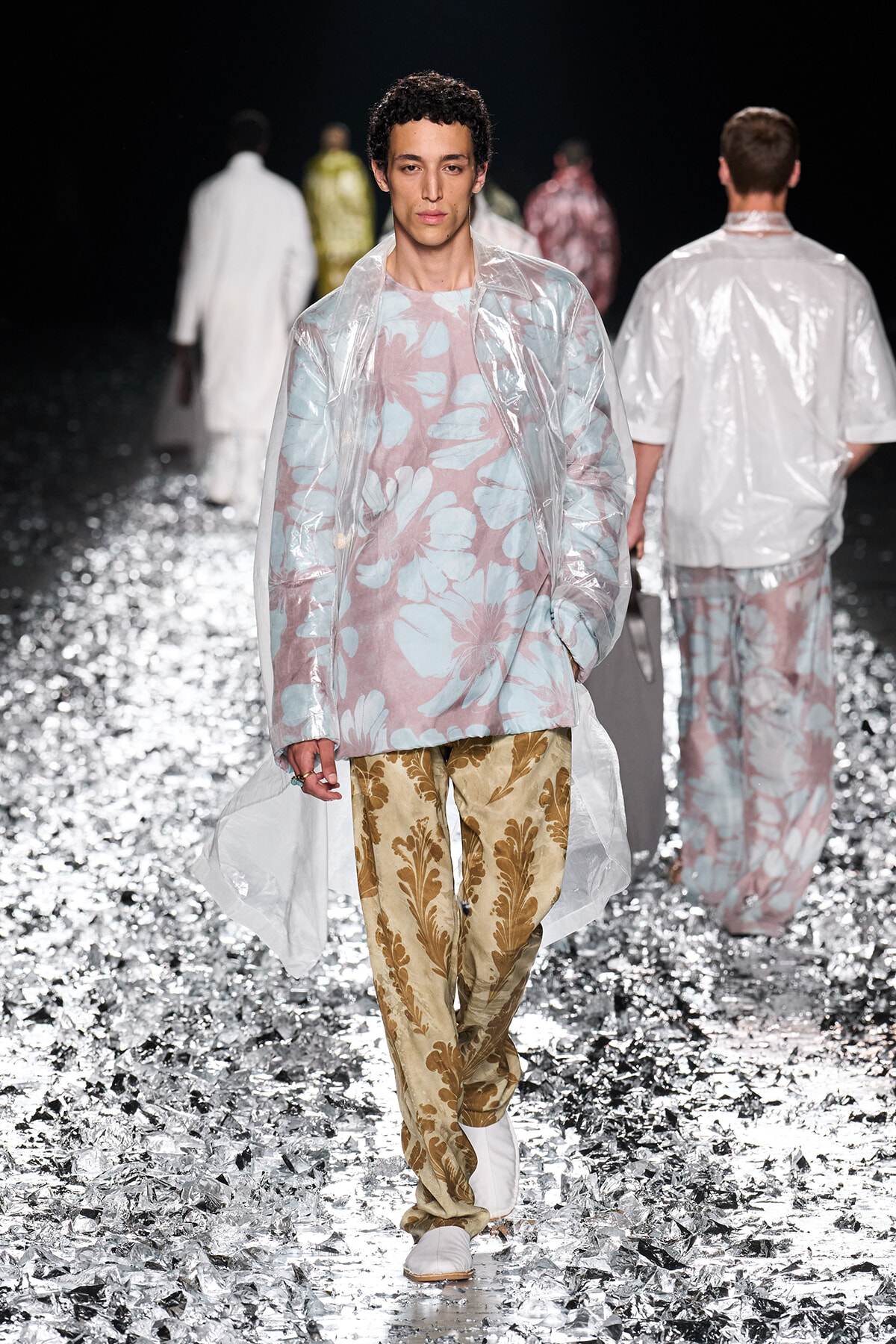 Model walks the runway in a clear plastic overcoat layered over a pastel pink shirt with a large blue floral print and gold brocade pants, while metallic confetti covers the floor.
