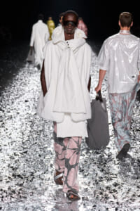 Model walking a runway in an oversized white hooded coat with pink floral-print pants and sandals, wearing dark sunglasses on a metallic confetti flooring.