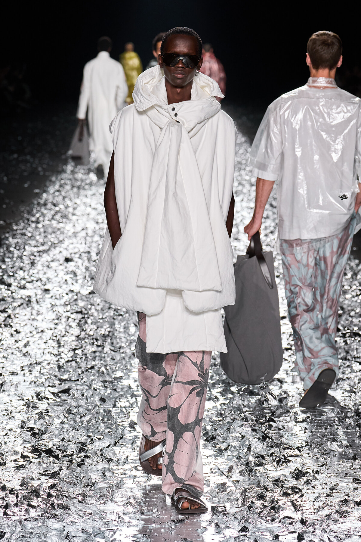 Model walking a runway in an oversized white hooded coat with pink floral-print pants and sandals, wearing dark sunglasses on a metallic confetti flooring.